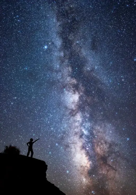 A sky full of stars with the denser milky way in the middle. A person in silhouette points to the stars.