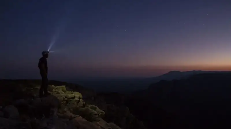 A hiker wearing a headlamp looks over the land as twilight sets in