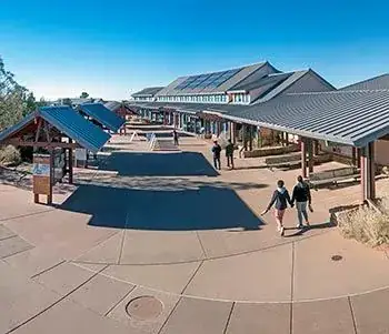 View down and across a concrete plaza; on the right are several information kiosks, on the right, a long narrow building with a gabled metal roof with banks of solar panels.