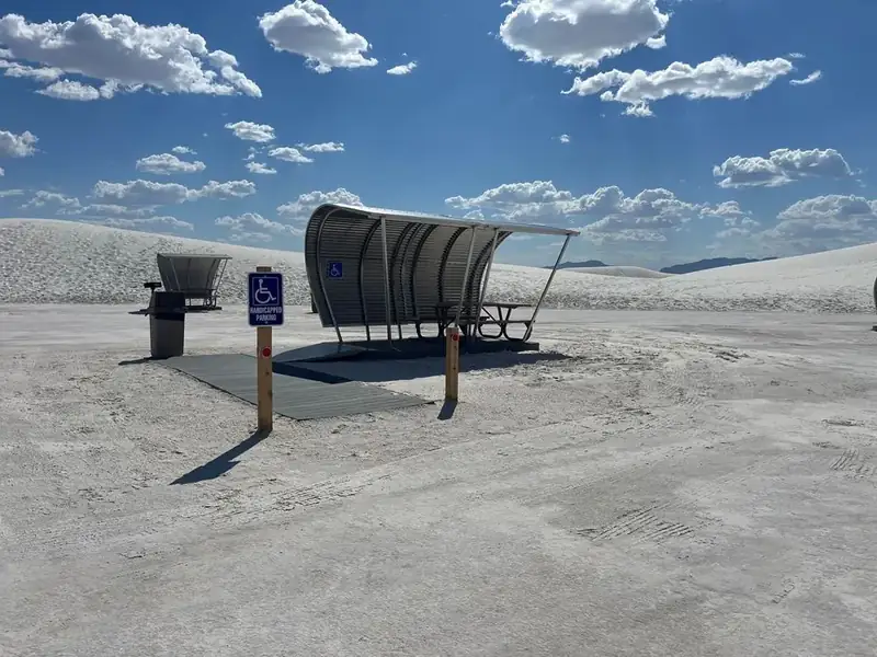 A curved metal structure covers a picnic table with an extended end. A ramp leads from the sand up to the concrete pad.